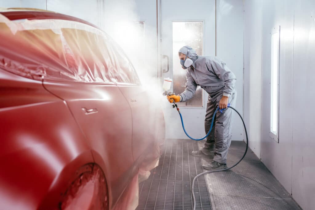 Peinture voiture professionnelle : carrossier repeint une auto Carrossier en combinaison intégrale et masque, peignant une voiture rouge avec un pistolet dans une cabine de peinture. Brouillard visible.