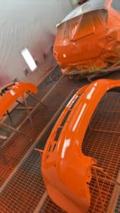 Atelier de peinture automobile avec une voiture orange bâchée et des pare-chocs fraîchement peints sur des grilles. Murs NDASA.