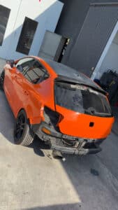 Voiture orange brillante dans un atelier, arrière sans pare-chocs ni feux. Le toit est noir et un sticker "MAZOUT GANG" est sur la lunette arrière.