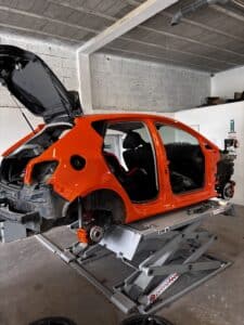Carrosserie orange de voiture dénudée sur un lift primetool 3200 kg en atelier. Intérieur de course, freins rouges. Panneau DAE.